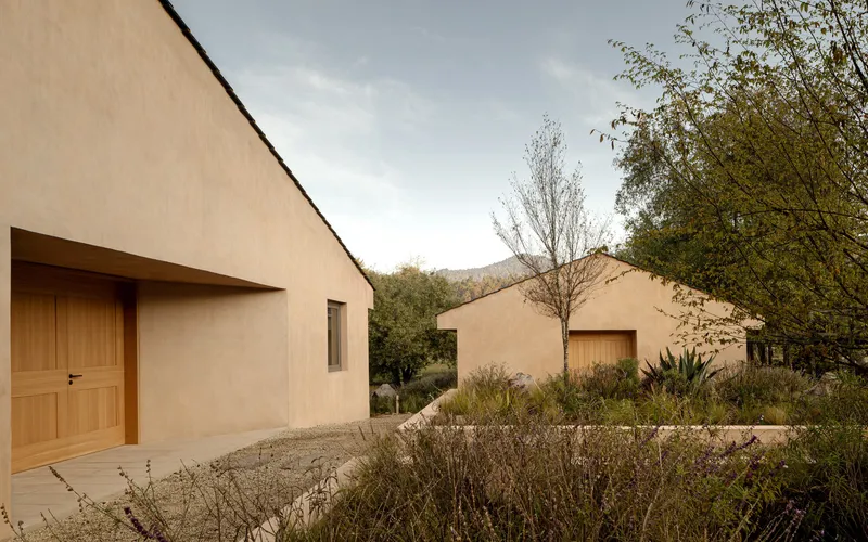 Gabled rammed earth volumes with timber doors opening onto a gravel courtyard with native plantings