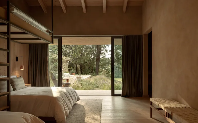 Bedroom with full-height glazing opening to a meadow under dappled morning sunlight