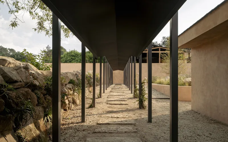 Covered walkway with black steel columns and overhead beams framing a gravel path flanked by boulders