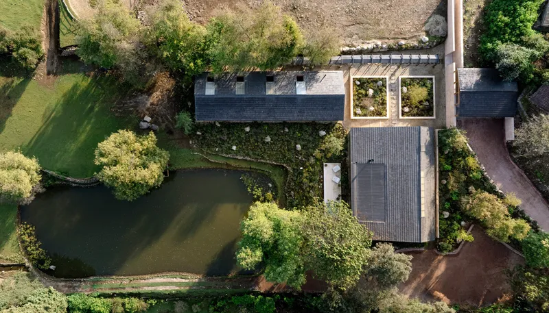 Aerial view of interconnected volumes with timber and slate roofs nestled between pond and lawn