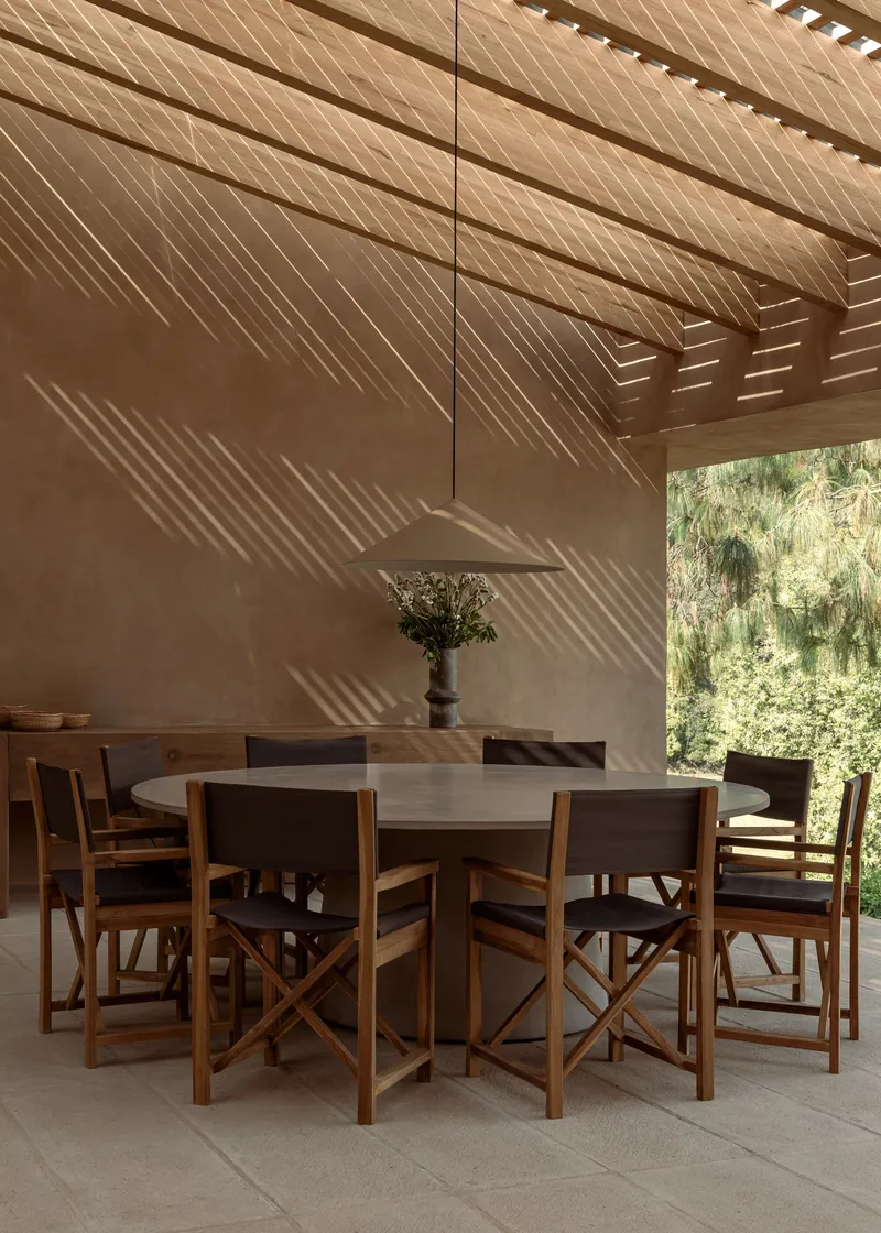 Dining area beneath slatted timber pergola casting diagonal shadow patterns on plaster wall
