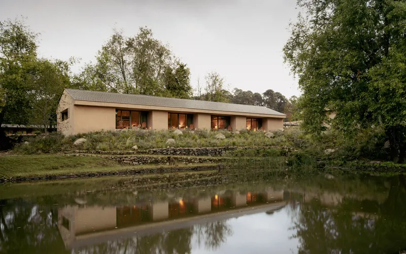 Long elevation of earth-toned building with regularly spaced window openings mirrored in pond water