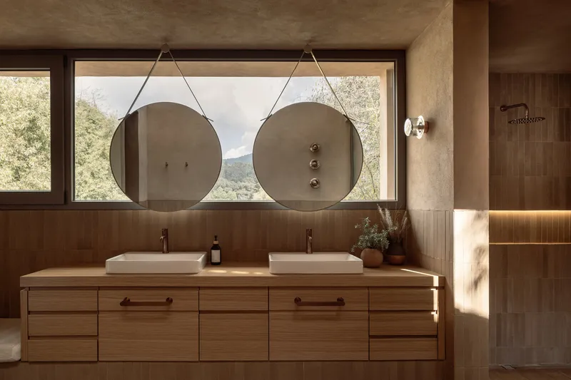 Double vanity in timber with round hanging mirrors framed by a horizontal window overlooking forested hills