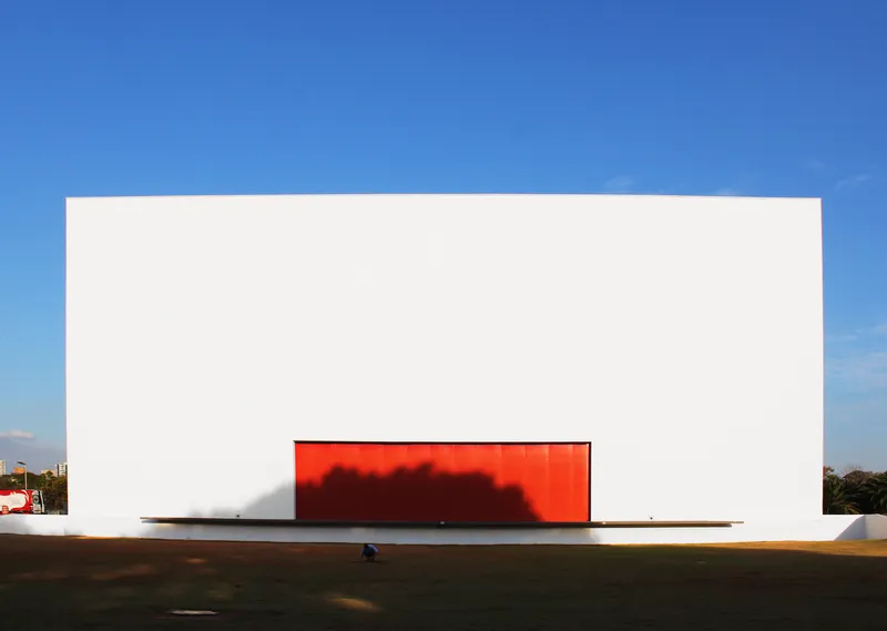 White cubic facade with a red backlit rectangular opening casting a shadow on the lawn at dusk