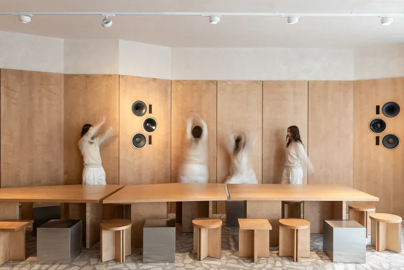 Curved plywood wall with circular speaker cutouts and blurred figures moving past a communal table