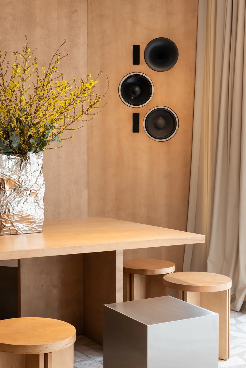 Corner dining area with a plywood table, cylindrical stools, and recessed circular speakers in the wall