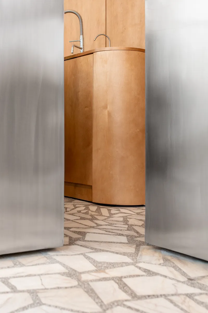 Curved plywood sink unit glimpsed through frosted glass panels above a terrazzo floor