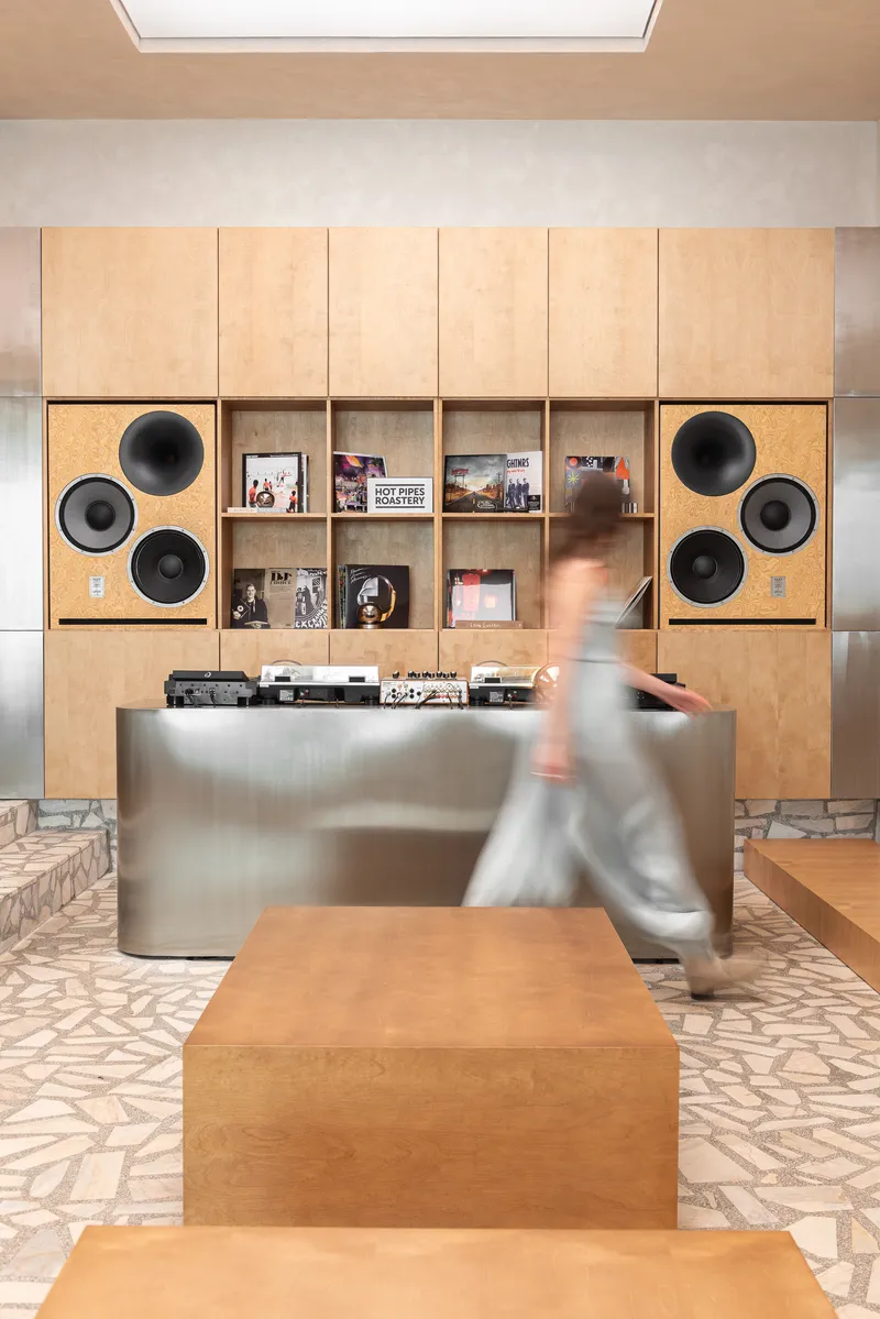 Plywood shelving wall with built-in circular speakers and a figure passing the brushed metal counter