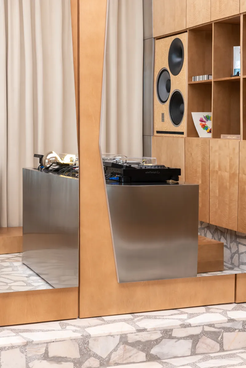 Detail of brushed metal DJ booth beside plywood wall with integrated speakers and record shelving