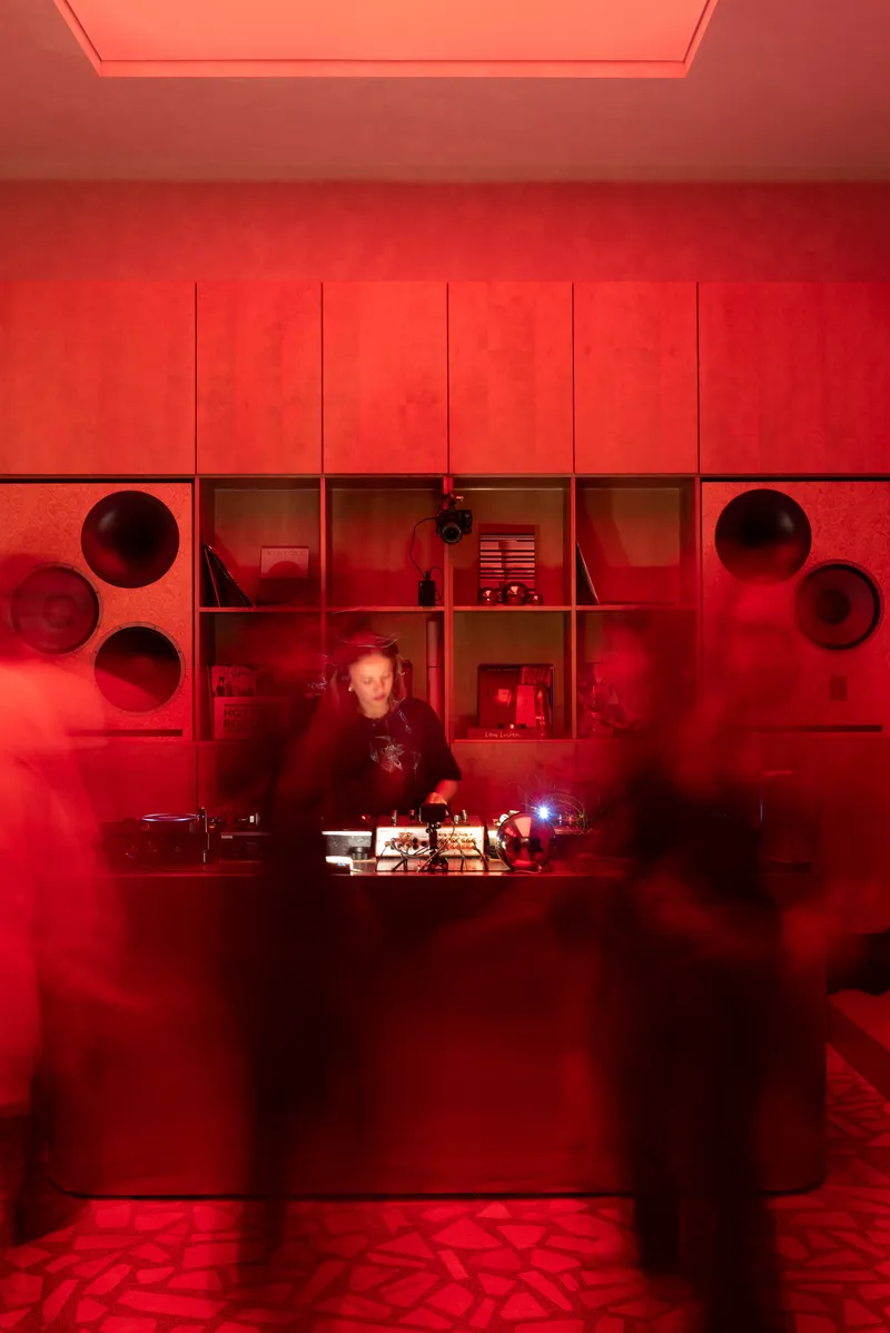 Red-lit service counter with circular openings and shelving units behind a figure working at dusk
