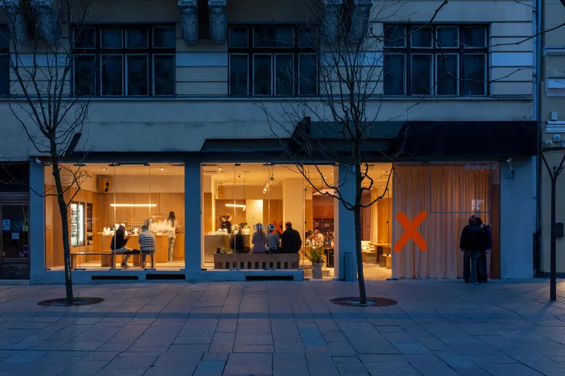 Street-level glazed shopfront with visitors inside at dusk, flanked by two bare winter trees