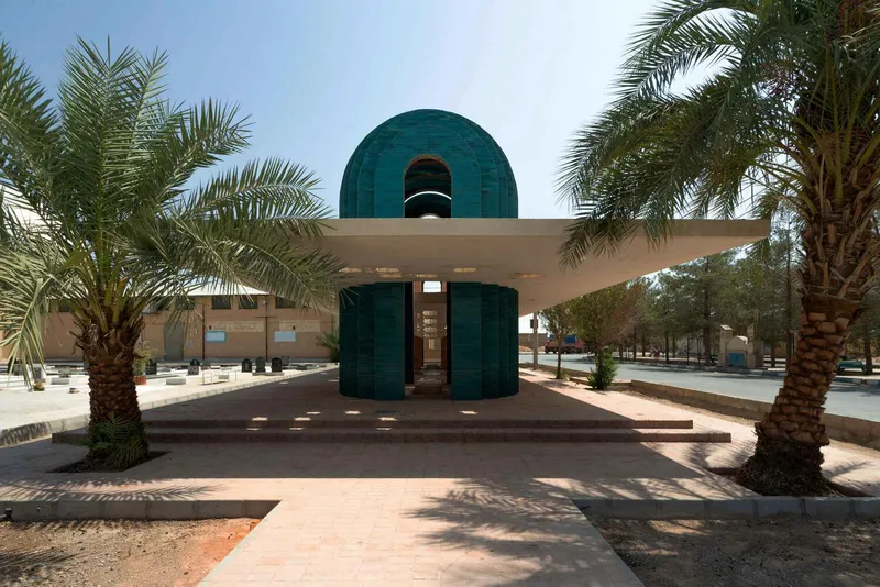 Front view of a cylindrical turquoise-tiled pavilion with horizontal roof canopy framed by date palms in daylight