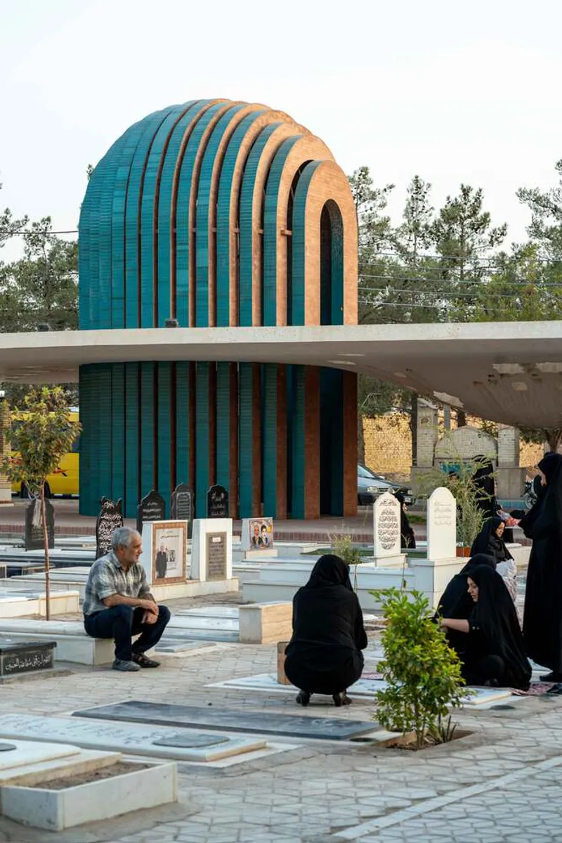 Ribbed barrel vault structure with alternating teal and terracotta bands above a paved cemetery courtyard