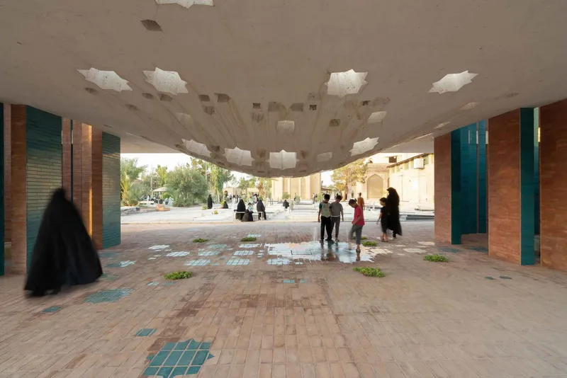 Brick-paved colonnade with turquoise columns and perforated ceiling casting shadows as visitors walk through the space
