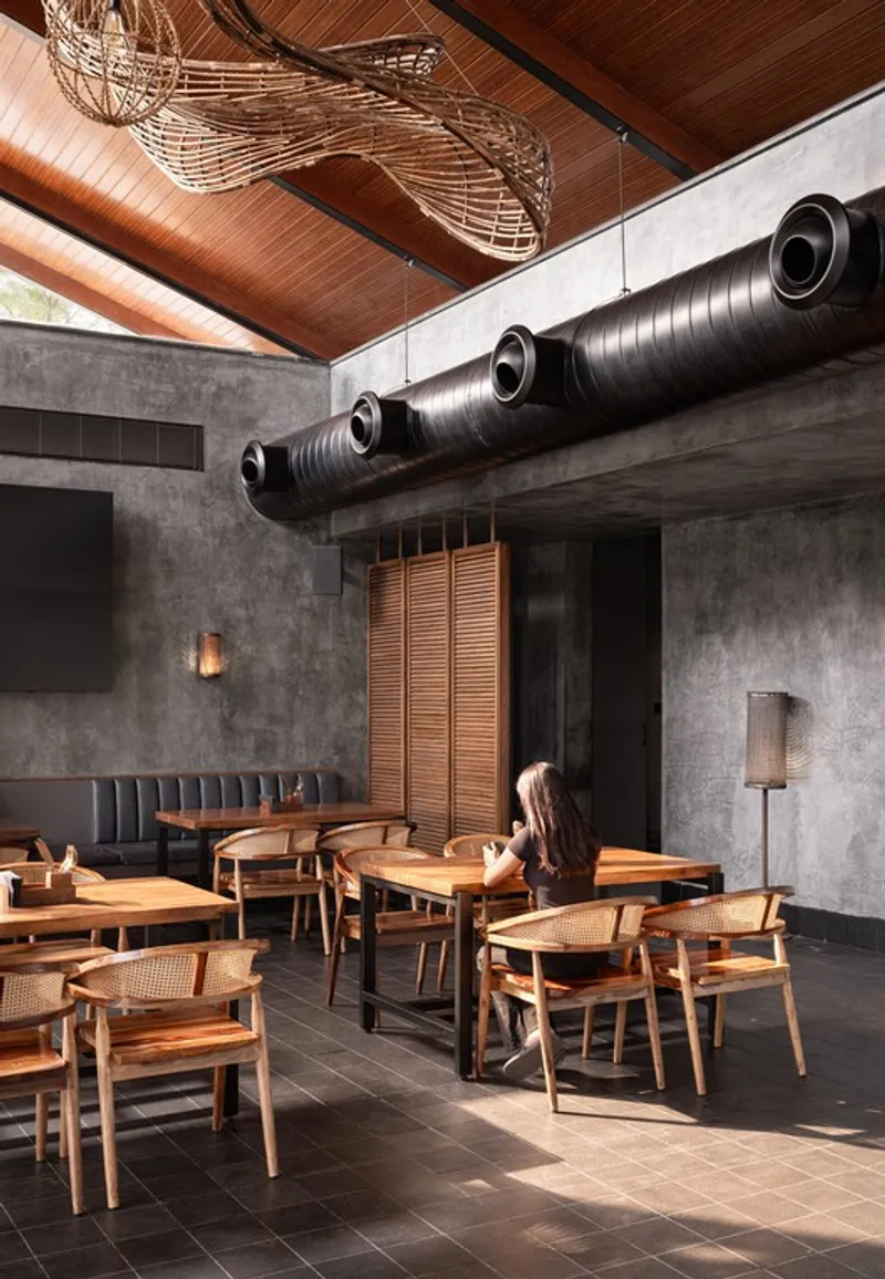 Dining area with exposed black ductwork, timber ceiling and sculptural woven pendant lights above concrete walls