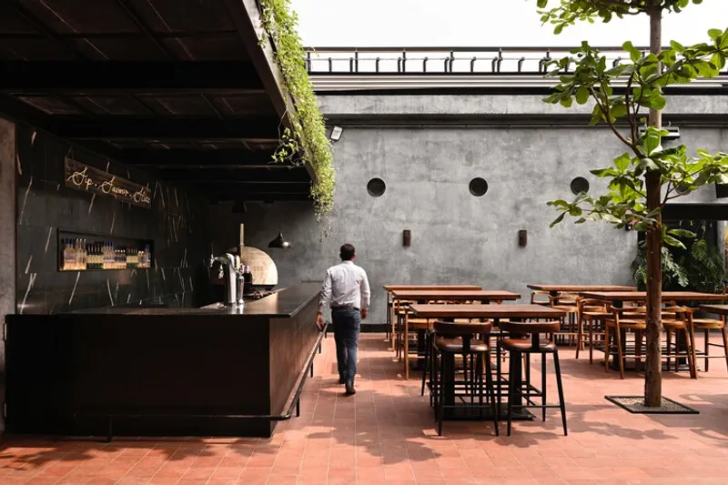 Courtyard terrace with metal bar counter, circular porthole windows in concrete wall and potted trees between tables