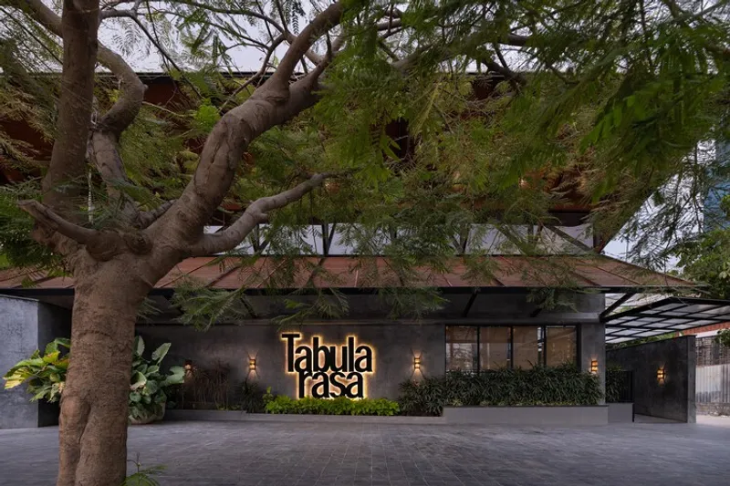 Street entrance facade with dark cladding and illuminated signage beneath a spreading tree canopy