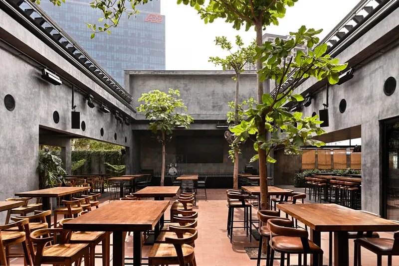 Open-air courtyard with wooden dining tables beneath trees surrounded by concrete walls with circular openings
