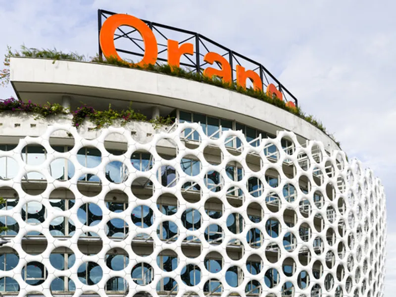 Curved facade with white perforated circular screen and orange signage on the roof terrace under cloudy skies