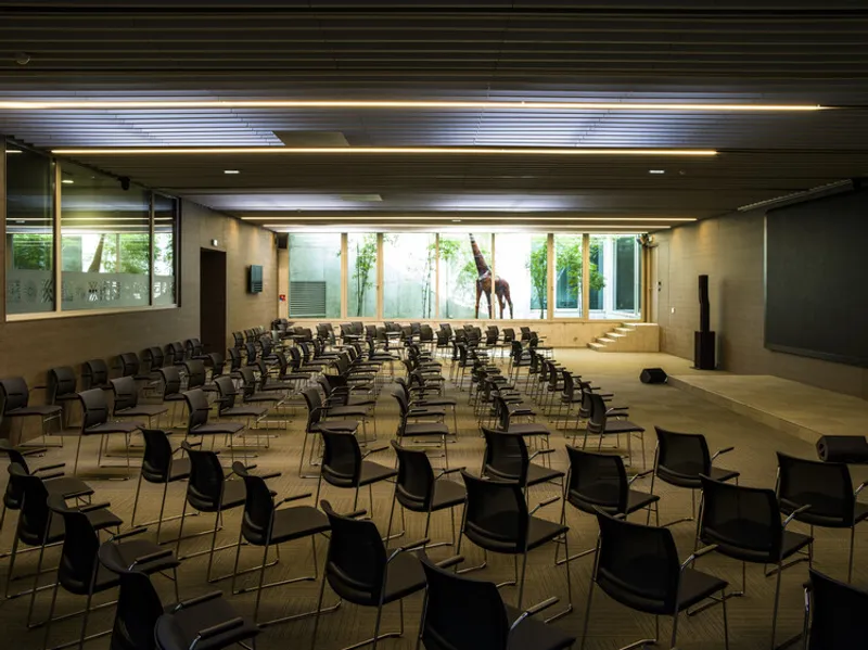 Assembly hall with rows of black chairs and timber slat ceiling facing a window wall with planted courtyard