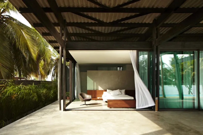 Covered outdoor terrace with corrugated metal roof, timber beams, and bedroom opening framed by palm trees