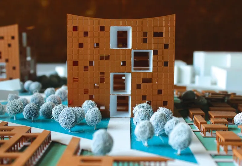 Detail of physical model facade with pixelated terracotta panels and stacked white window openings beside cotton ball trees