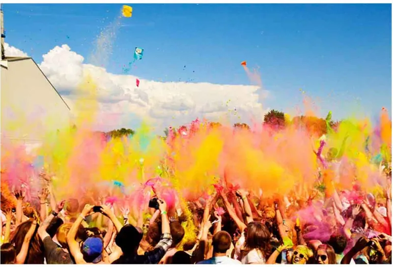 Festival crowd throwing colored powder into the air beneath a blue sky with scattered clouds and kites