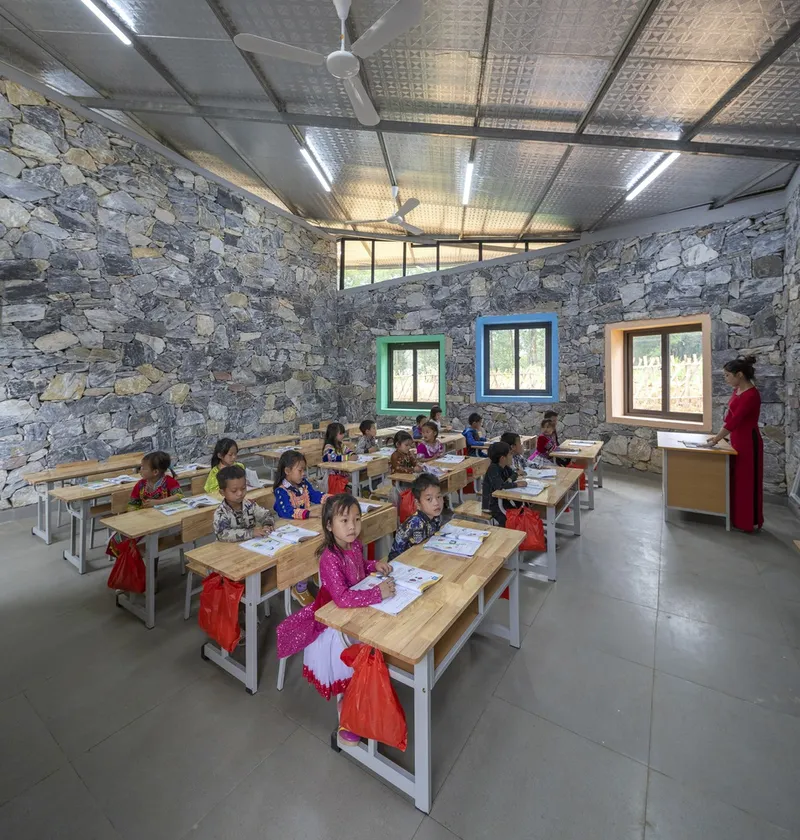 Classroom interior with stone walls, corrugated metal ceiling with exposed beams, and rows of timber desks with children studying