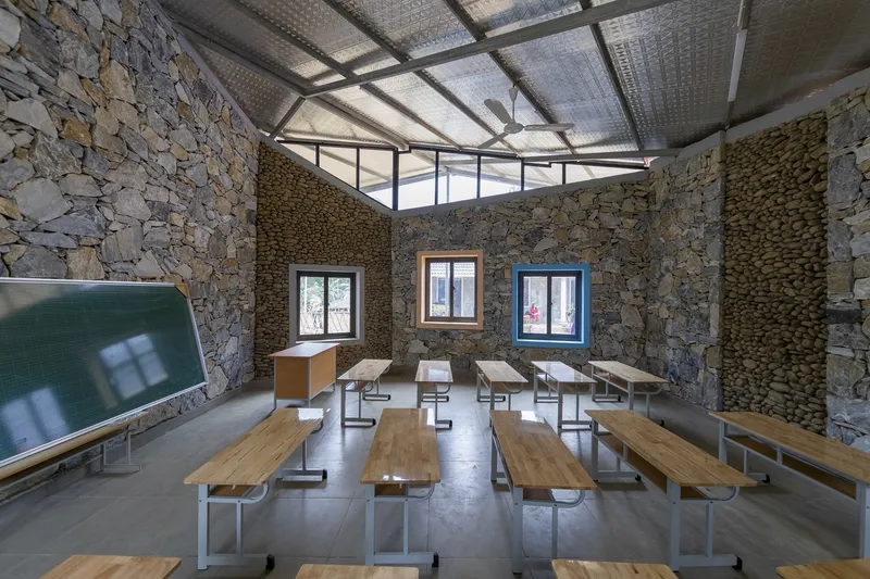 Stone-walled classroom with three colored window frames and timber benches under a gabled metal roof