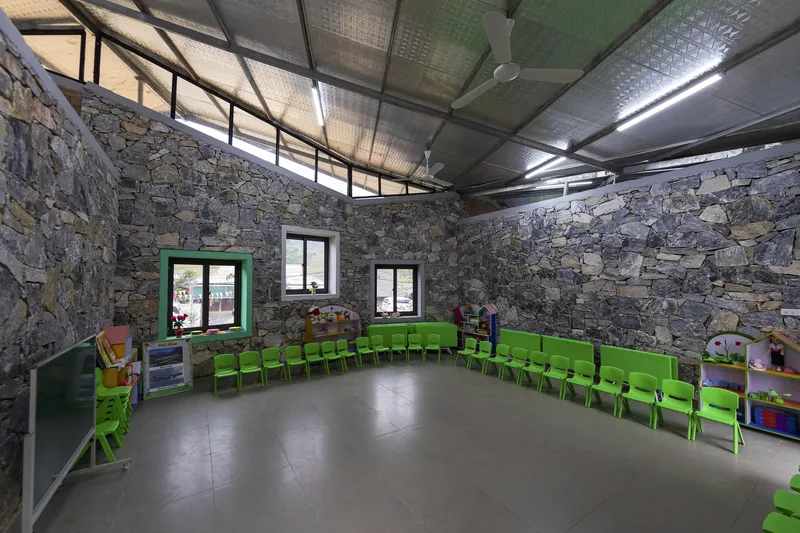 Classroom interior with exposed stone walls, corrugated metal ceiling, and rows of green plastic chairs