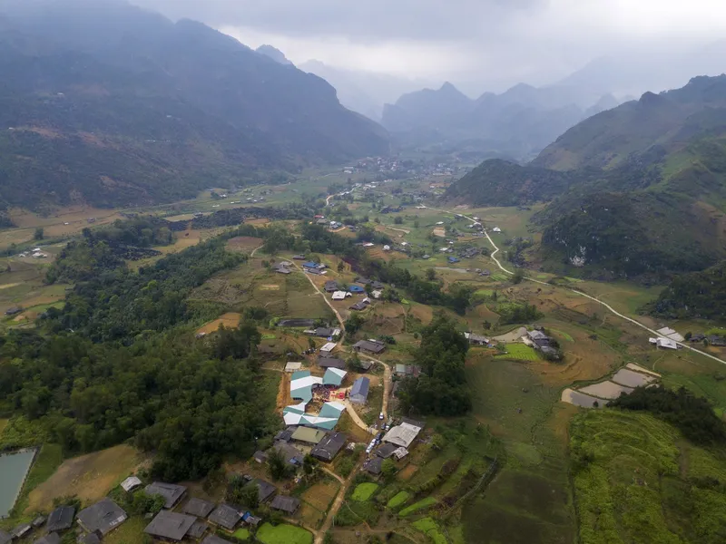 Aerial view of scattered buildings with metal roofs set among terraced fields and forested hills in mist