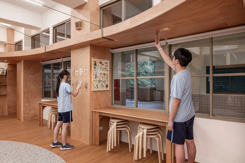 Two students at a counter with pegboard and stacked stools beneath a timber-clad mezzanine level