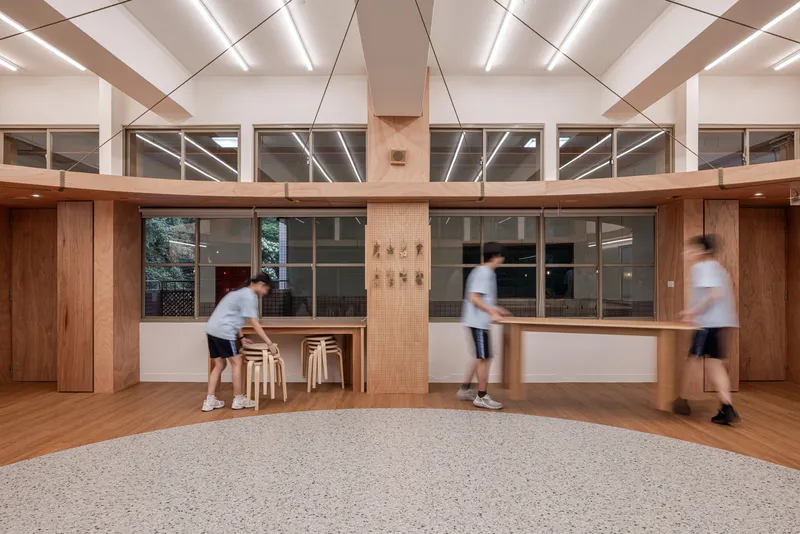 Double-height common area with timber central counter, diagonal ceiling grid and people in motion
