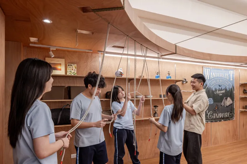 Interior room with curved plywood ceiling beams and pegboard wall where students gather around suspended rope swings