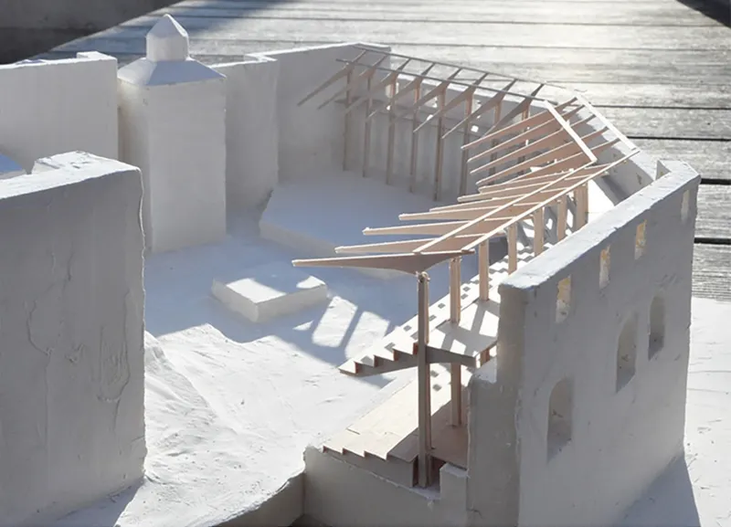 Physical model showing exposed timber roof framing rising above white masonry walls in sunlight