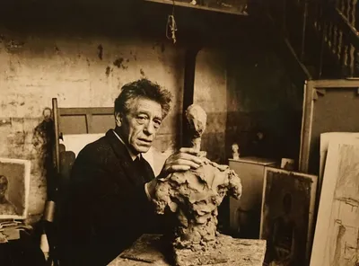 This is a sepia-toned photograph of an artist in his studio, working on a clay sculpture. The artist