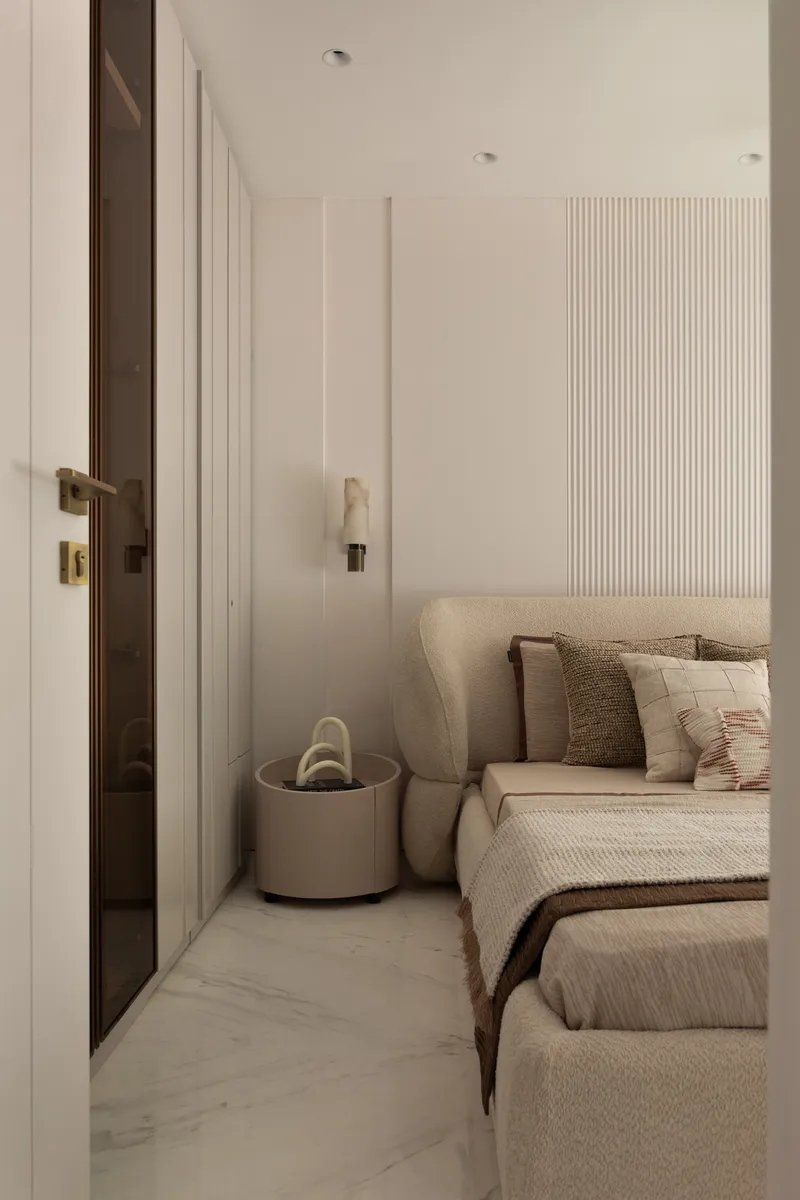A side view of the bouclé bed alongside a tinted brass-framed glass door and an alabaster wall sconce.