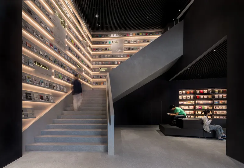 Concrete staircase ascending past illuminated bookshelves with readers seated below at dusk
