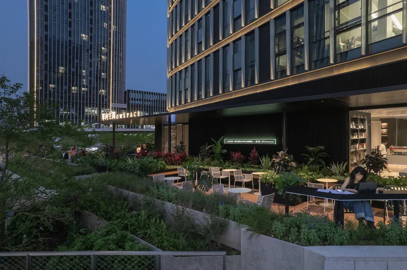 Outdoor seating area with planted beds and visitors gathered at twilight beneath tower facades