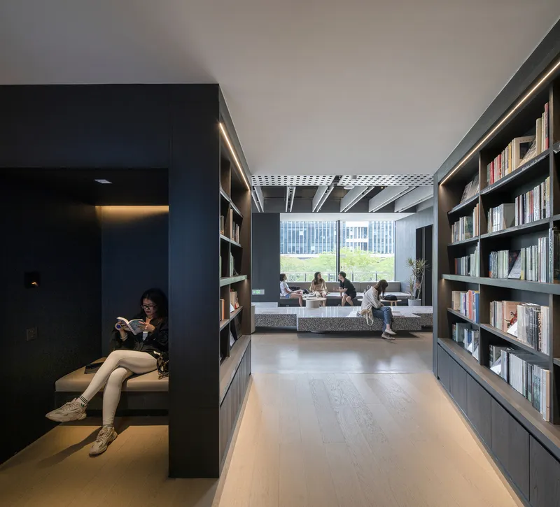 Reading alcove with built-in seating and a visitor reading, opening onto a sunlit space with bookshelves