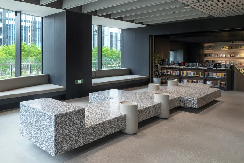 Terrazzo benches with cylindrical stools arranged beneath floor-to-ceiling windows overlooking tree-lined street