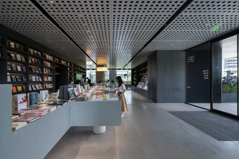 Bookstore interior with perforated metal ceiling, display tables, and a visitor browsing near full-height shelving