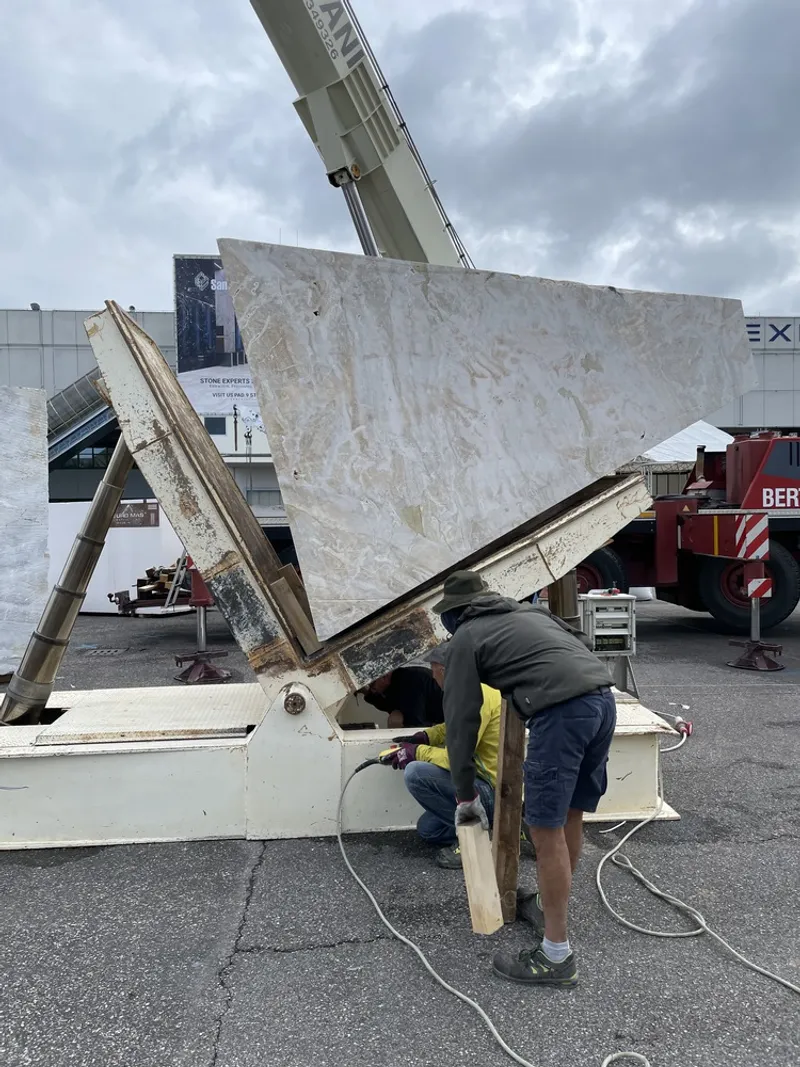 Worker securing large marble slab suspended by crane on a cloudy day outside an exhibition center