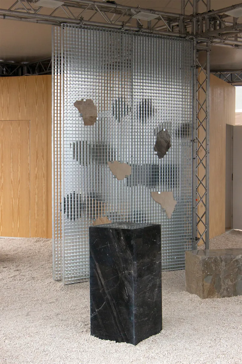 Translucent gridded screen suspended from metal truss above a black marble plinth on gravel flooring