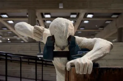 The image showcases a sculpture of a human figure seemingly trapped in a vise, set against the backd