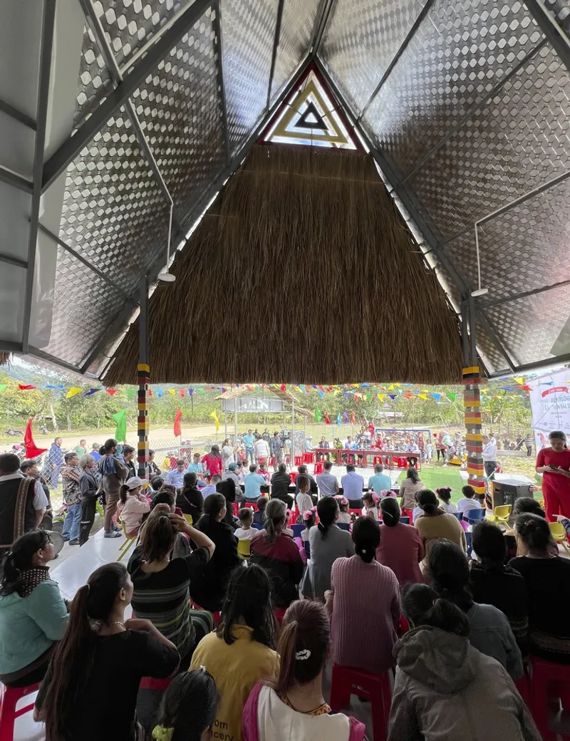 View from under the thatched pyramidal roof toward an outdoor gathering with seated audience and perforated metal ceiling