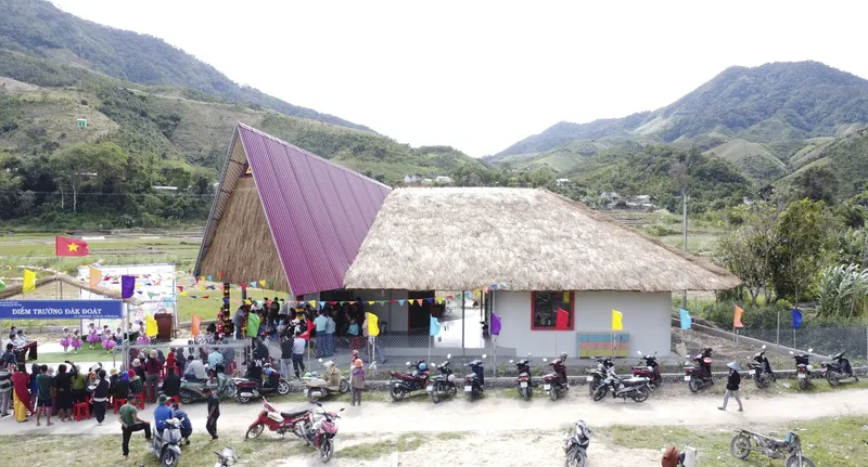 Front elevation showing the split pavilion with thatched and pink corrugated roofs during a community gathering with motorcycles parked