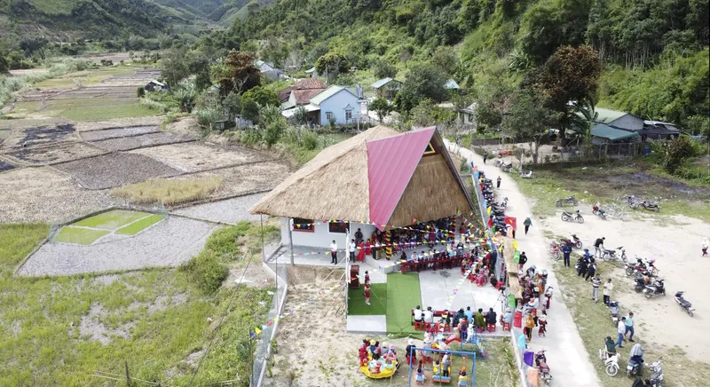 Aerial view of the thatched-roof structure with triangular profile amid rice paddies and gathered villagers