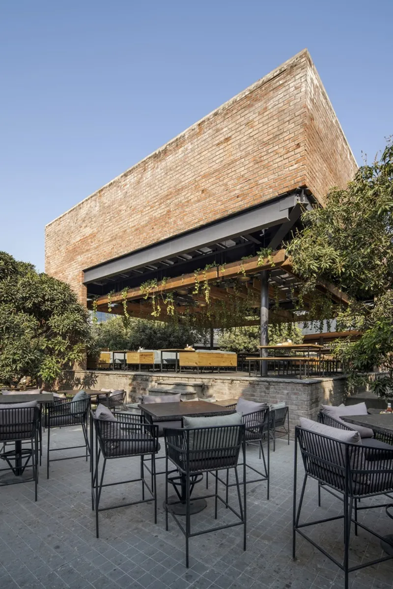 Outdoor seating area beneath a cantilevered brick volume and steel pergola surrounded by mature trees