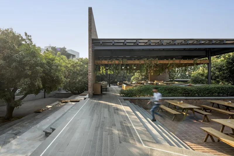 Covered terrace with steel frame and dappled sunlight casting shadows across stone paving and picnic tables